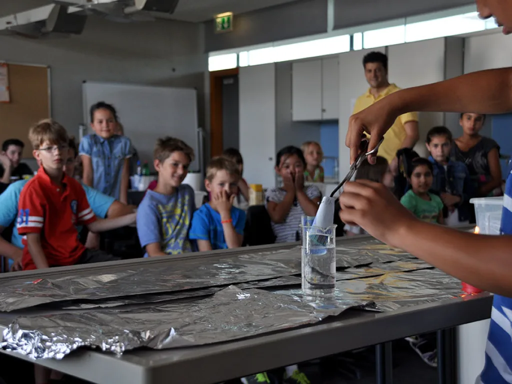 Kinder stehen um einen Tisch und verfolgen ein Experiment.